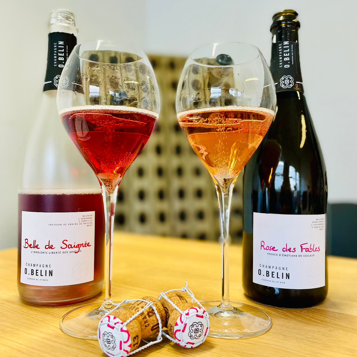 2 flasker champagne på bord med 2 glas fyldt med roséchampagne, 2 propper og pupitre i baggrunden. Champagne O. Belin, Belle de Saigné og Rose des Fables