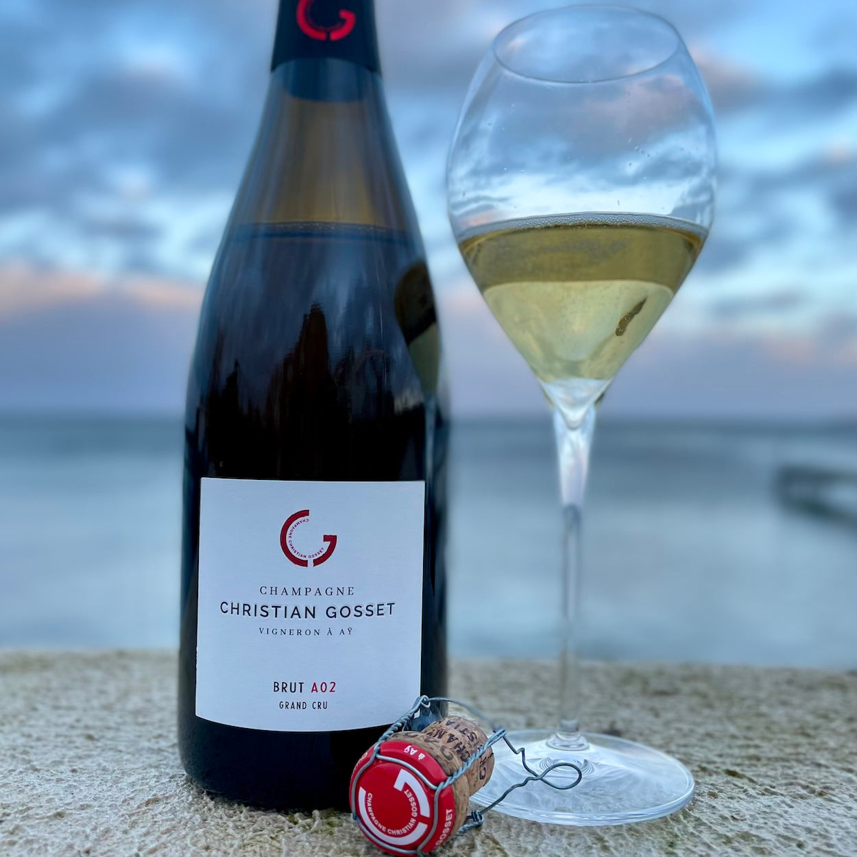 Champagne flaske og fyldt champagneglas står med udsigt til have og himmel, champagneprop, Champagne Christian Gosset Brut A02 Grand Cru