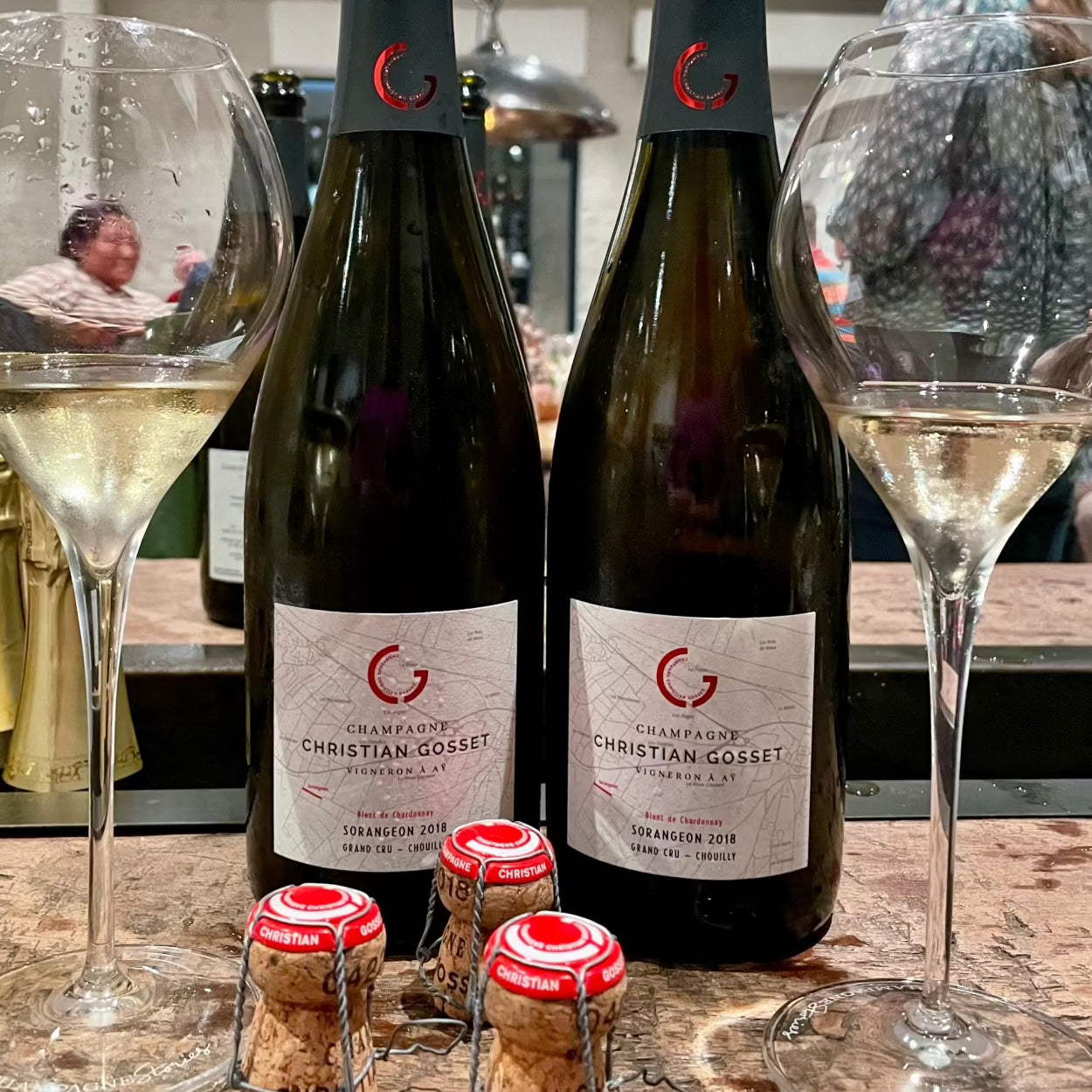 2 flaske champagne på bord med 2 champagneglas og 3 champagnepropper, mennesker i baggrunden, Champagneflaske på bord med champagneglas foran fyldt med rosé champagne, Champagne Christian Gosset, Sorangeon 2018, Blanc de Chardonnay, Grand Cru