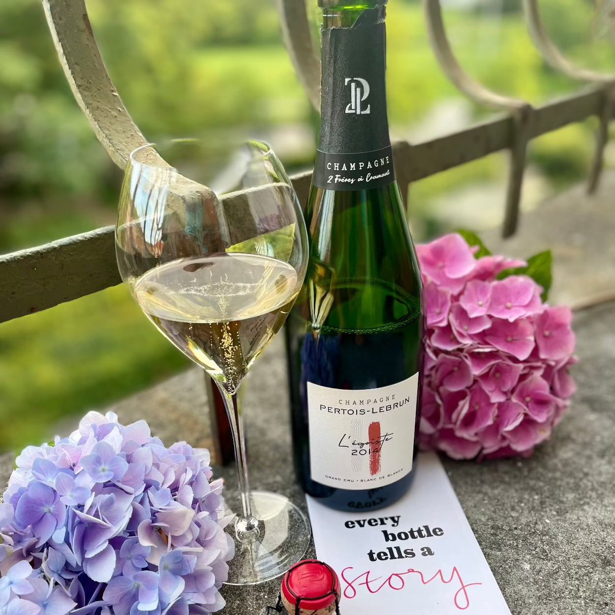 Champagne flaske på altan, champagneglas fyldt med champagne til venstre for champagneflasken, bobler i glasset, hortensia blå og lyserød på hver side, champagneprop med rød kapsel, postkort med teksten^every bottle tells a story", Champagne Pertois-Lebrun, L'egoïste 2014, Grand Cru, årgangschampagne