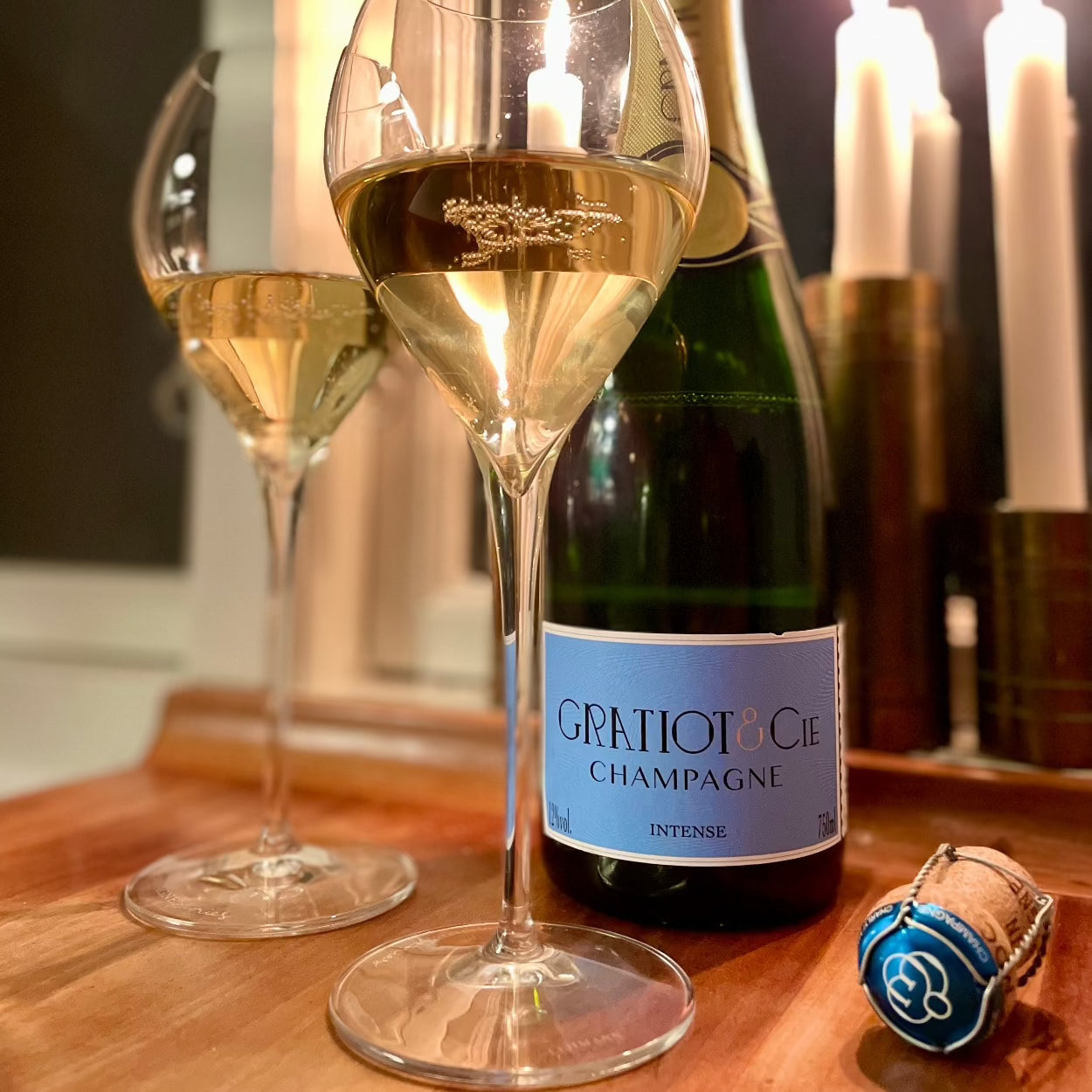 1 flaske champagne på bord med 2 glas med champagne fra Champagne Gratiot & Cie, Intense, blå etikette, tændte sterinlys i baggrunden og champagne proppen ved flasken, blå med logo