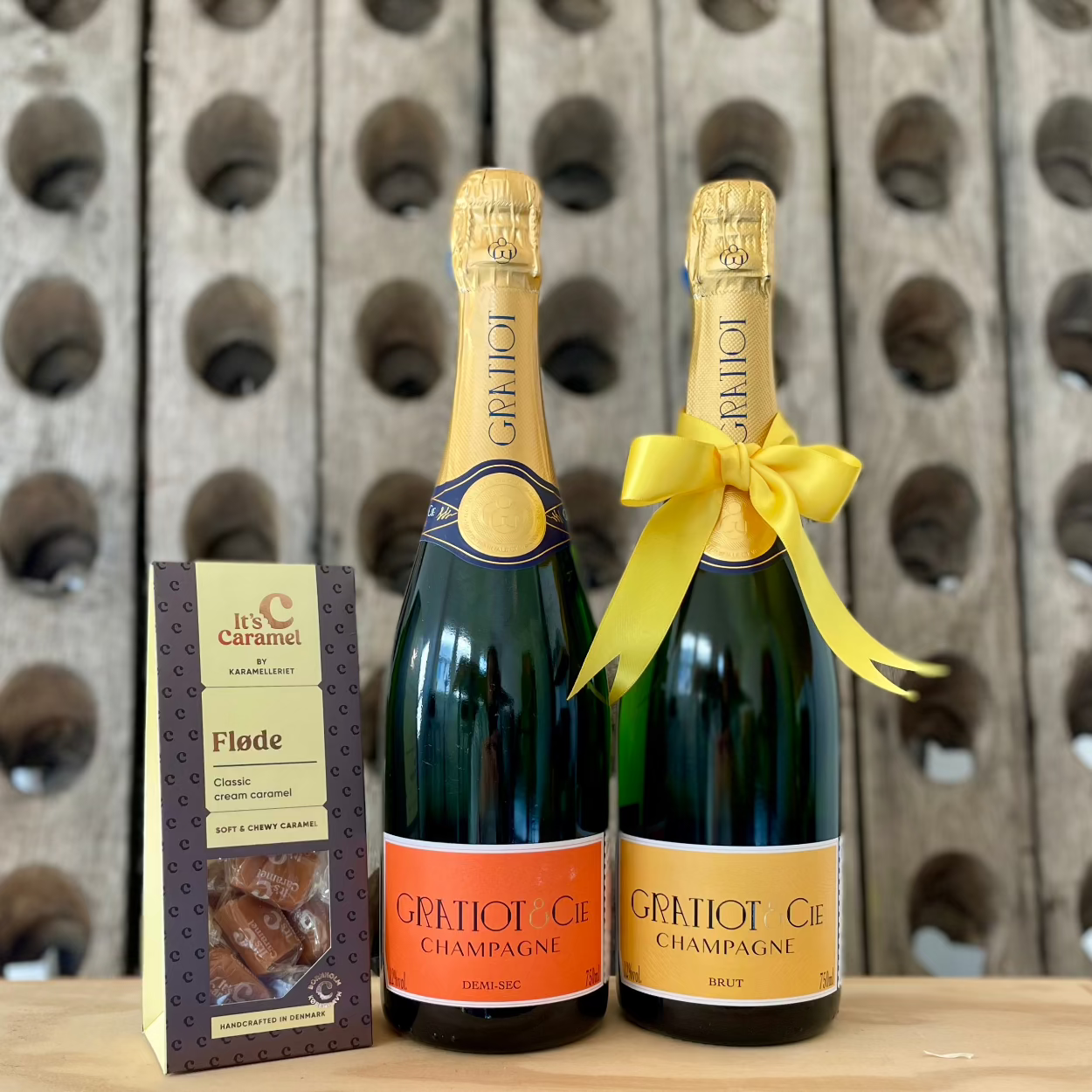 2 flasker champagne og 1 pakke flødekarameller med pupitre i baggrund. Champagne Gratiot & Cie, Almanach no. 1, Demi-sec og Brut, sidstnævnte med en gul sløjfe om halsen