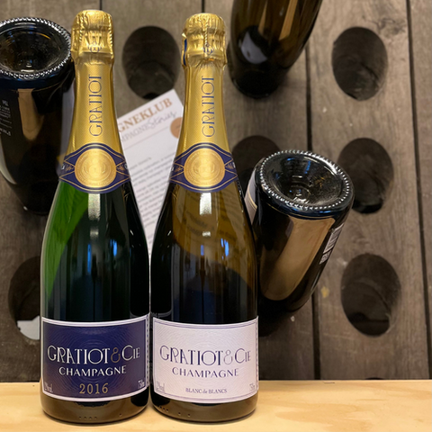 2 flasker champagne fra Champagne Gratiot & Cie, Champagne Almanach nr. 4, Milésime 2016 og Champagne Almanach nr. 6, Blanc de Blancs. Bagrunden er en pupitre med nogle flasker og en klubflyer