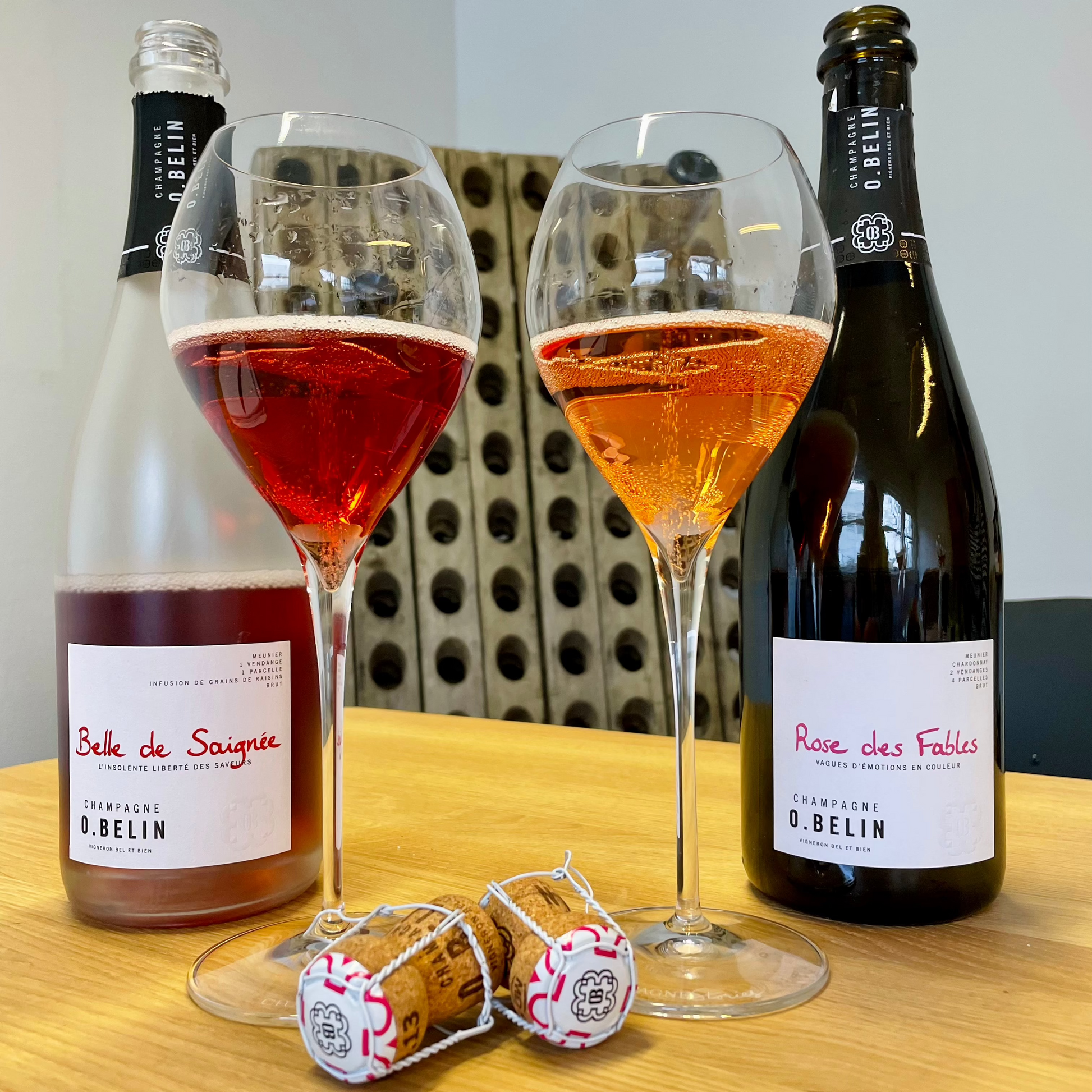 2 flasker roséchampagne, 2 champagneglas fyldt med roséchampagne og 2 propper. Champagne O. Belin, Belle de Saignée og Rose des Fables. Pupitre i baggrunden