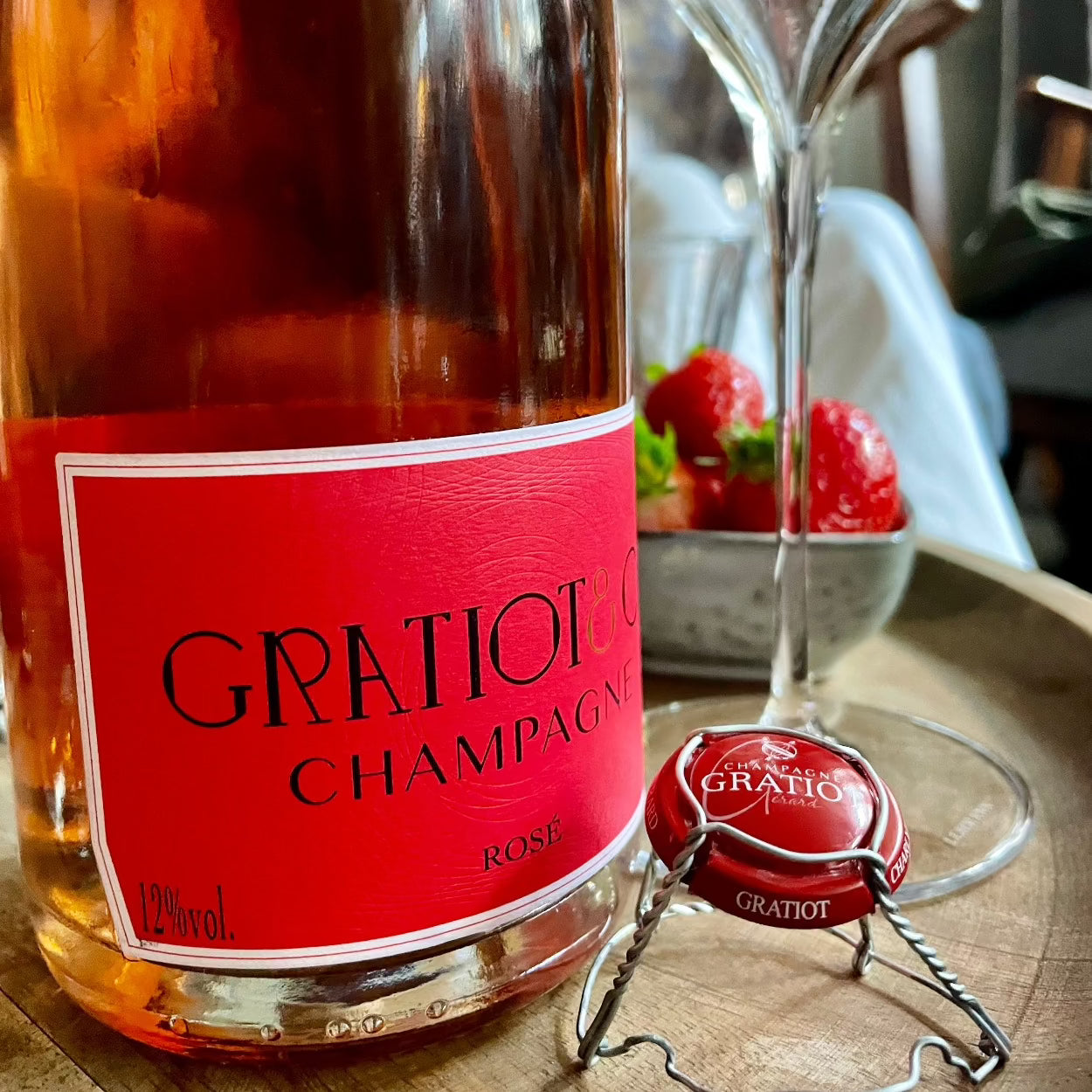 tæt på champagne flaske med rosé champagne, rød etikette, champagne fra Gratiot & Cie, Rosé, muslet og kapsel ved siden af flasken, glas, jordbær i baggrunden