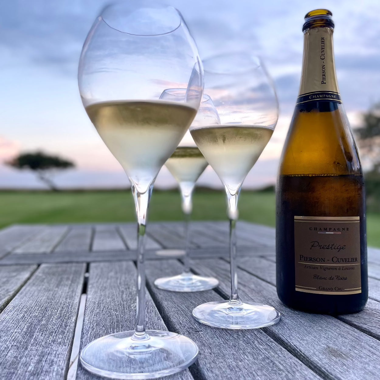 1 flaske champagne fra Champagne Pierson-Cuvelier, Prestige, Grand Cru, på et havenord, ved siden af 3 Lehmann Grand Jamesse champagneglas med champagne, uskarp solnedgangshimmel i baggrunden